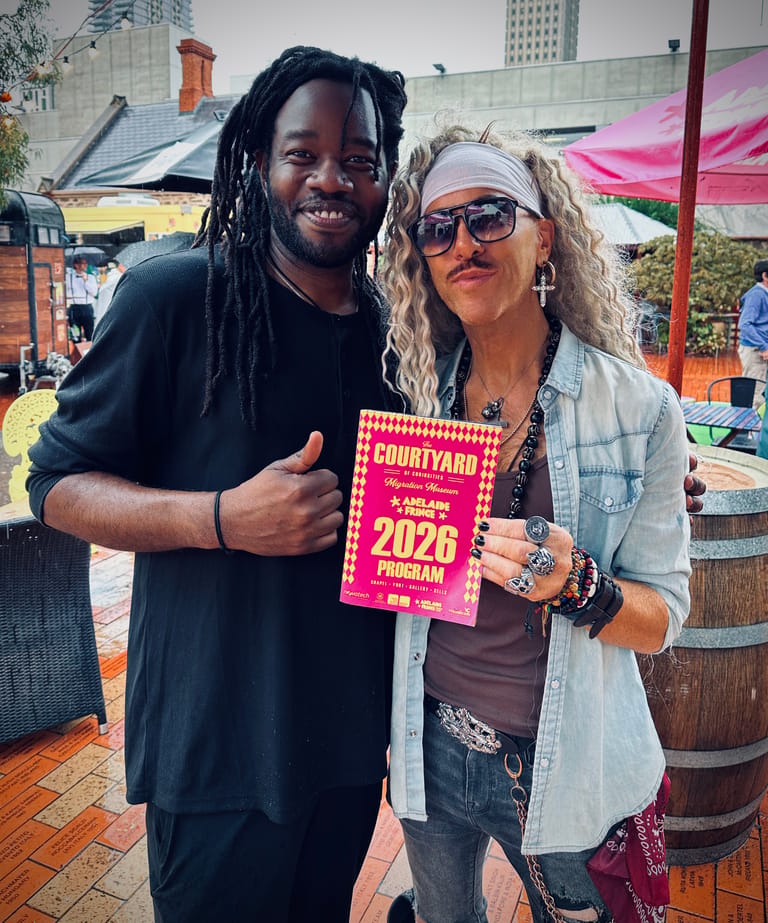 Two people smile at an outdoor Adelaide Fringe venue, one holding a bright pink 2026 programme.