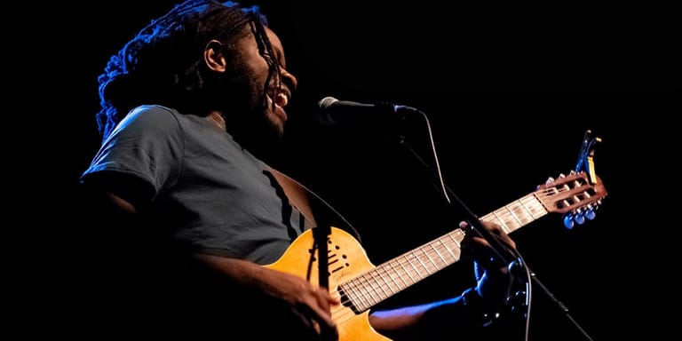 Musician in dark shirt playing acoustic guitar at microphone under blue stage lighting.
