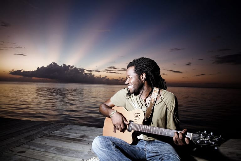 Musician with dreadlocks playing acoustic guitar on a wooden deck at sunset over water.