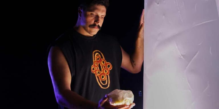 Performer in black sleeveless shirt with orange graphic holds glowing orb under blue stage lighting.