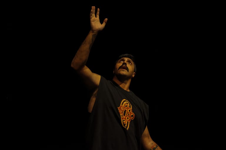 Performer in black shirt with raised arm against dark background during stage performance.