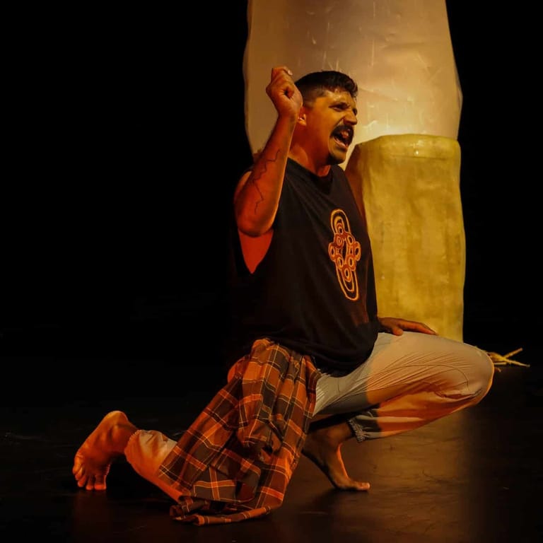 Performer kneels on stage with fist raised, wearing black shirt and checked pants, illuminated by warm stage lighting.