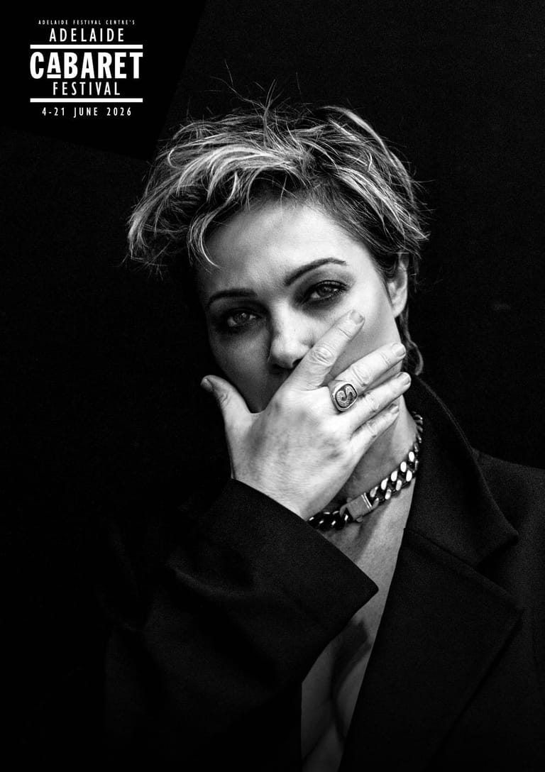 Black and white portrait of a performer in a dark suit with short textured hair, hand raised to face, wearing rings and a chain bracelet against a dark background.