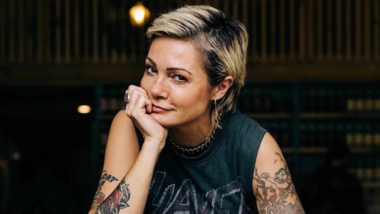 Performer with short blonde hair and tattoos on their arm, resting their chin on their hand in a contemplative pose.