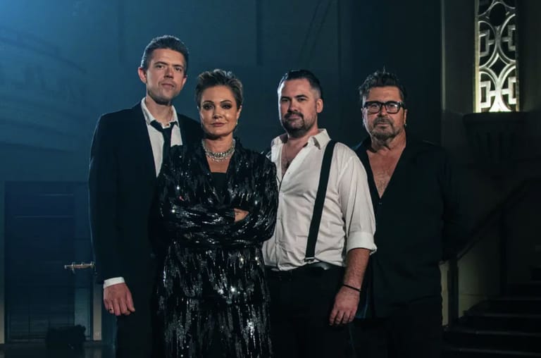 Four performers stand together against a blue-lit backdrop in cabaret-style formal attire.