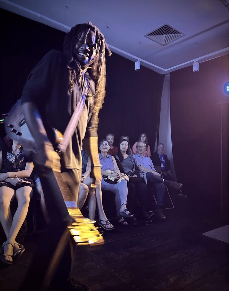 Performer with dreadlocks in dramatic gold lighting moves across stage while seated audience members watch from the side.