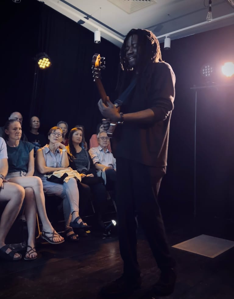 Performer with long dreadlocks holds a microphone on stage, addressing seated audience members under warm stage lighting.