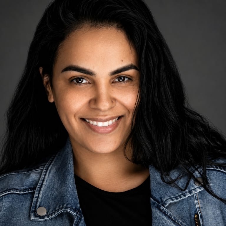 Person with long dark hair and a bright smile wearing a denim jacket over a black shirt, photographed against a neutral grey background.