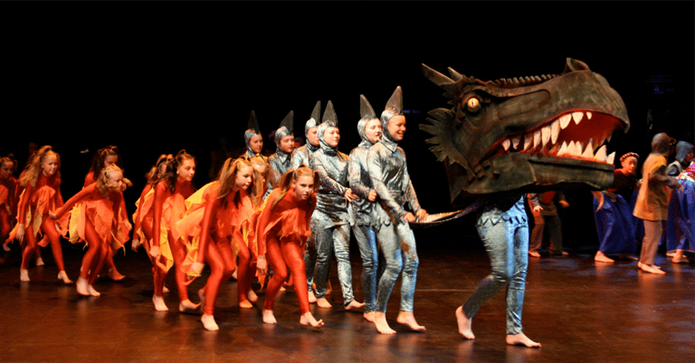 Children in costume dance alongside a large dragon puppet on a theatre stage.