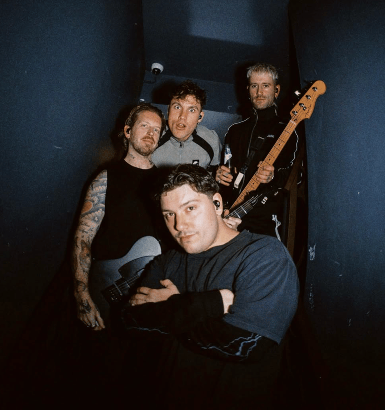Four musicians pose together in dim blue-toned lighting, one holding a bass guitar.