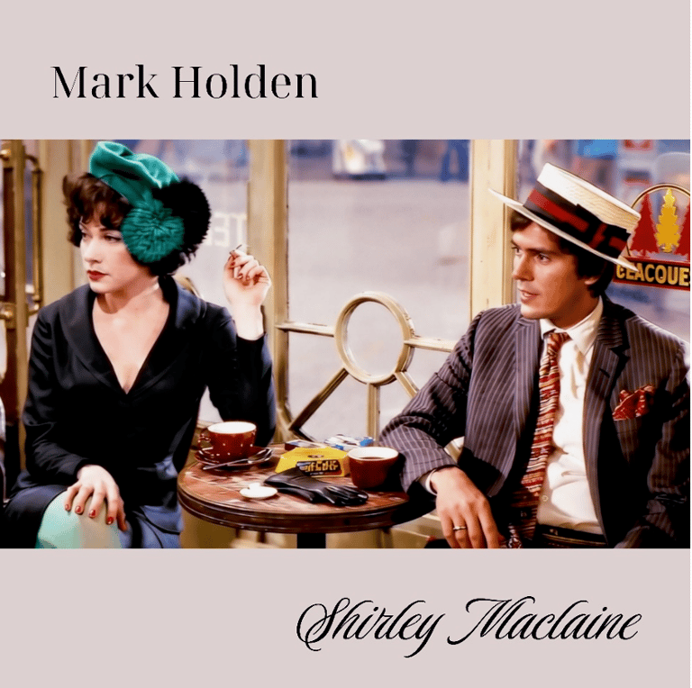 Two performers in vintage 1920s attire sit at a table with theatrical props during a period scene.