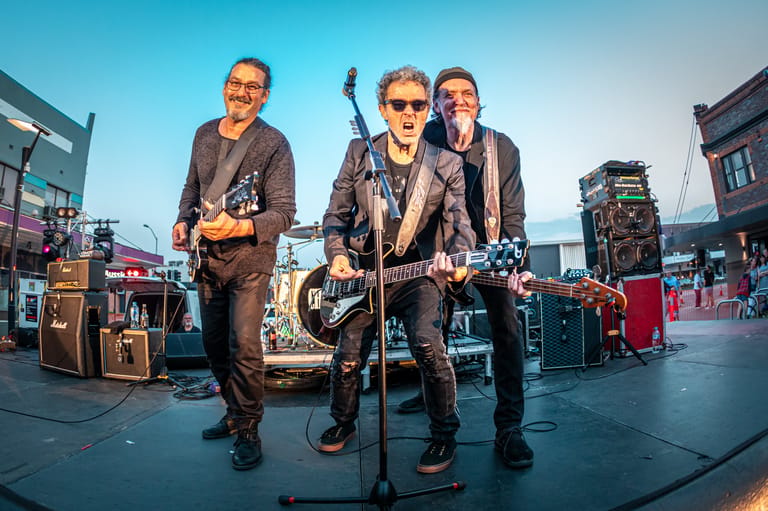Three musicians perform with guitars on an outdoor stage at dusk, with city buildings visible in the background.