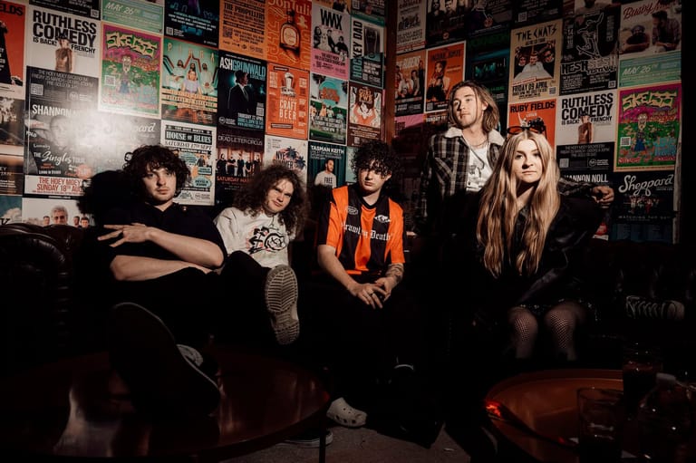 Five band members pose in a darkly lit room covered with music posters and memorabilia.