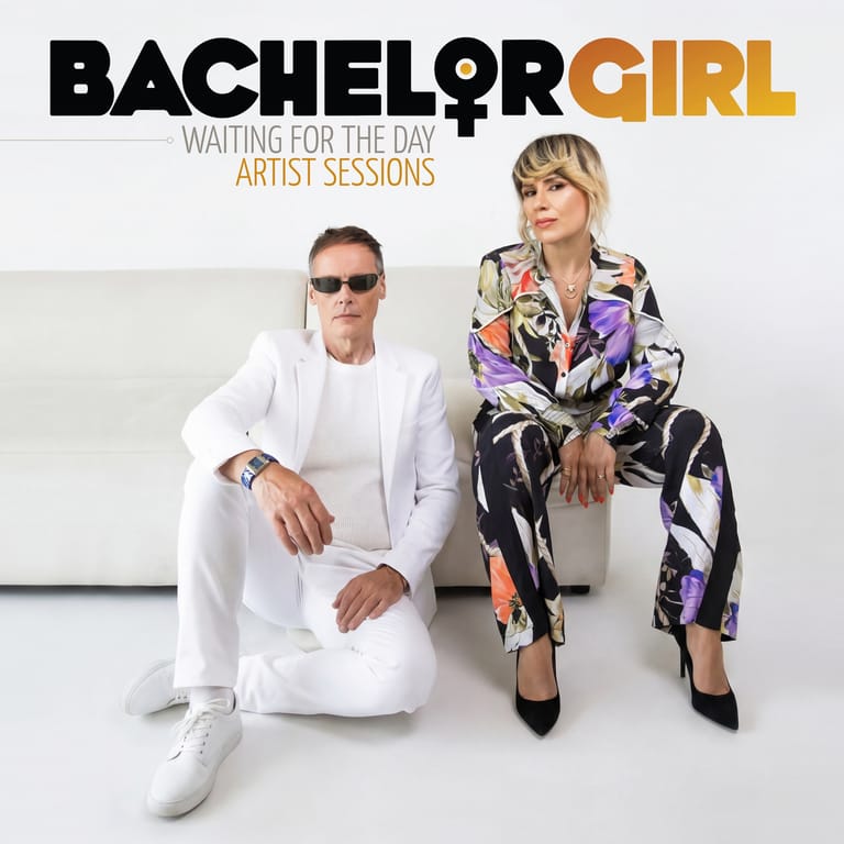 Two musicians sit against a white backdrop, one wearing all white and sunglasses, the other in a patterned jacket with blonde fringe.