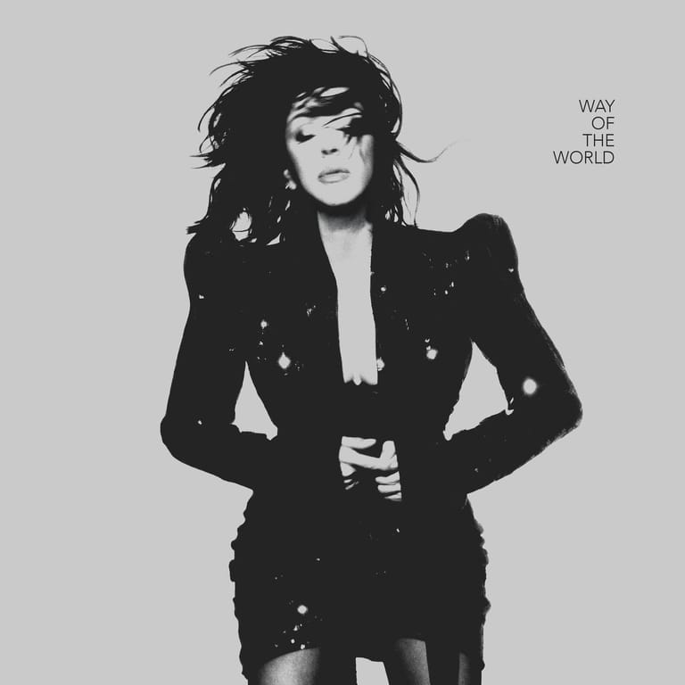 Person in black embellished jacket with tousled dark hair against grey background, hands on hips.