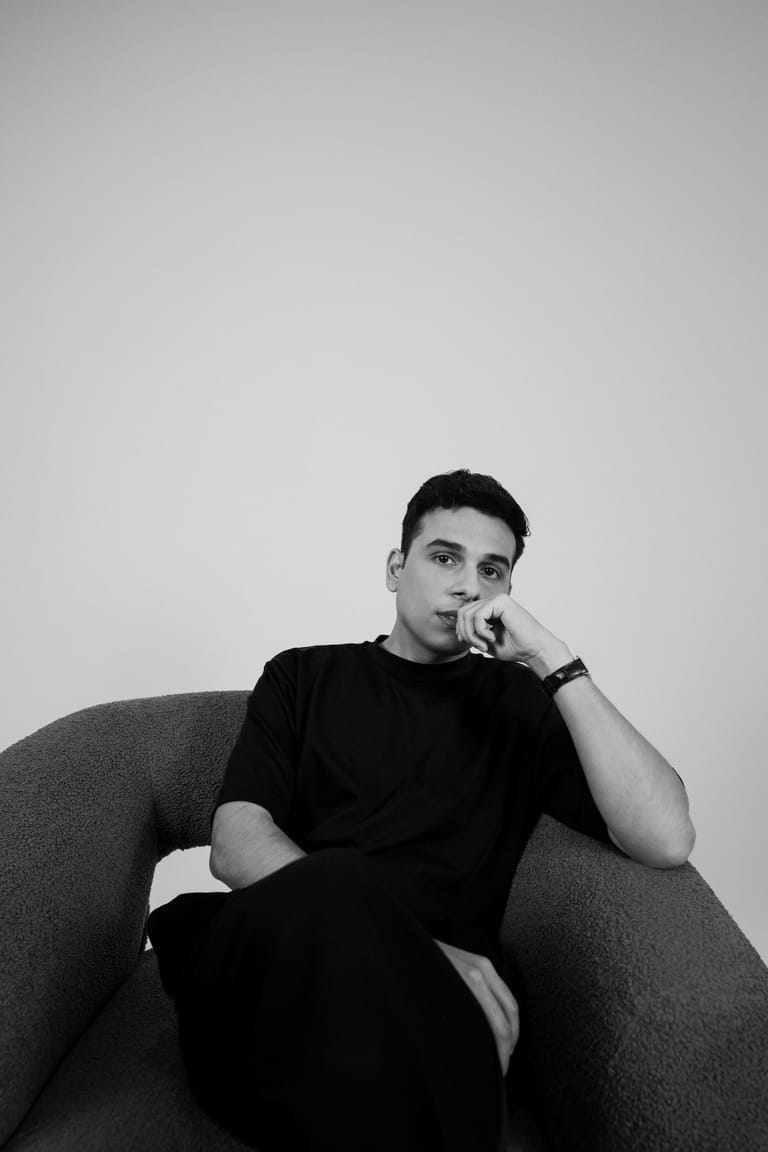 Man in black shirt seated in grey armchair, hand to face in contemplative pose.
