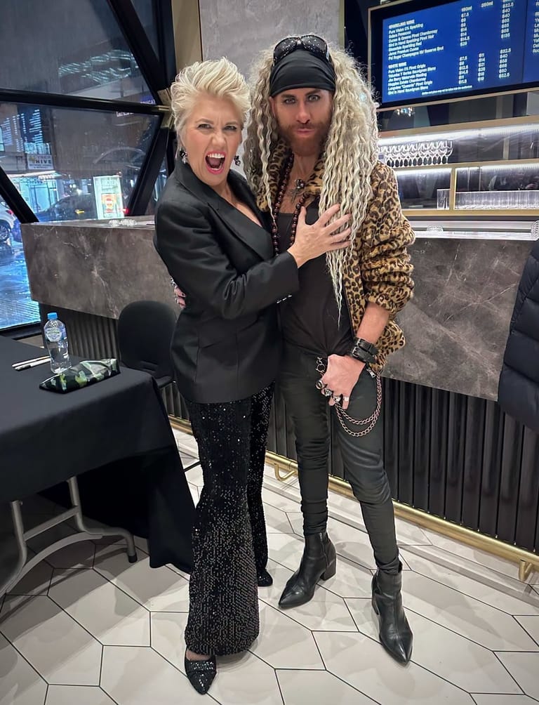 Two performers pose together backstage, one in a black blazer and the other wearing a leopard-print jacket and long blonde hair.