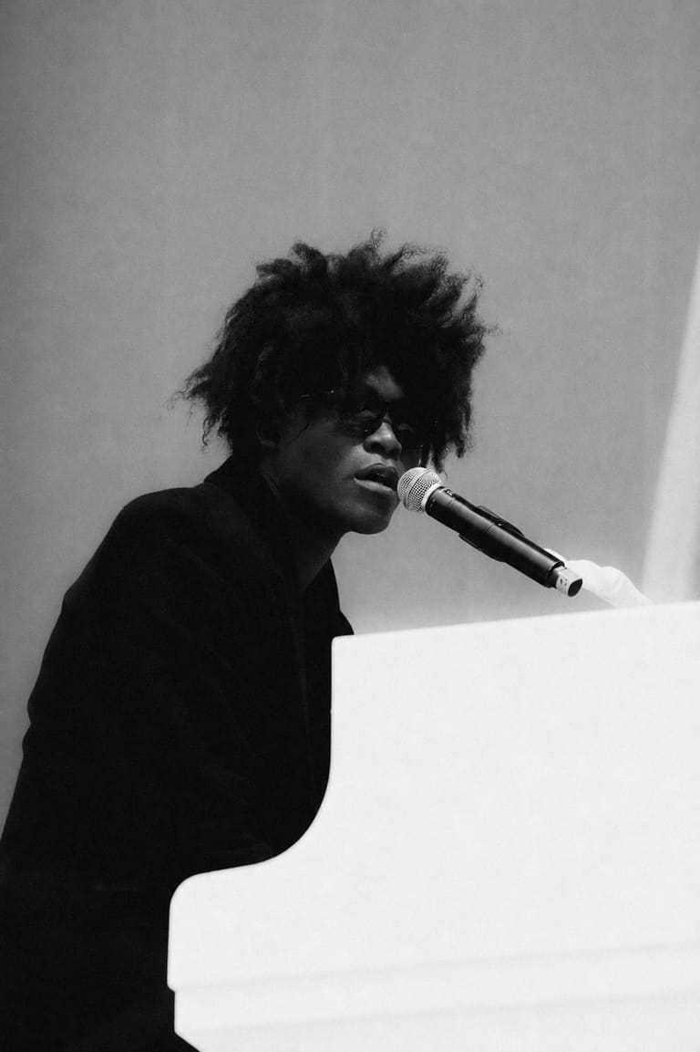 Musician with voluminous dark hair singing into microphone at white piano in black and white portrait.