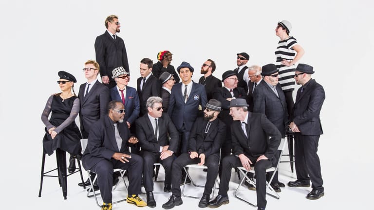 Large ensemble of performers in formal black suits and striped dress arranged on white chairs against a plain background.