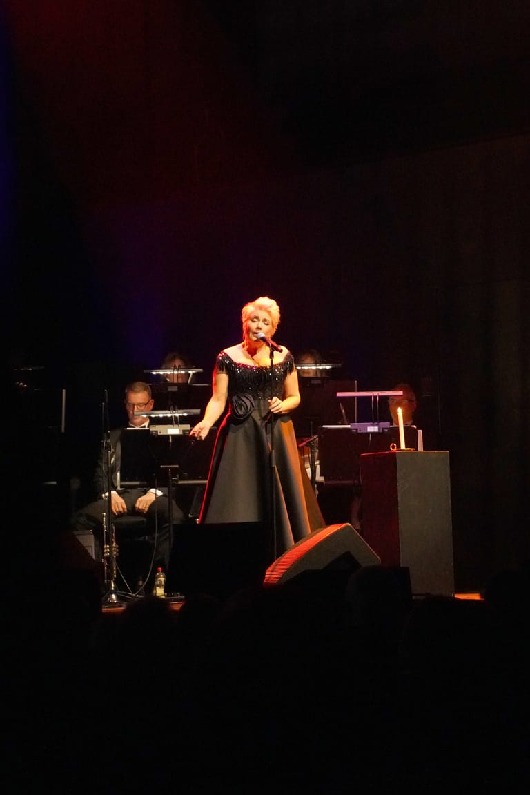 Female performer in black dress and red boots singing at a microphone on a dimly lit stage.