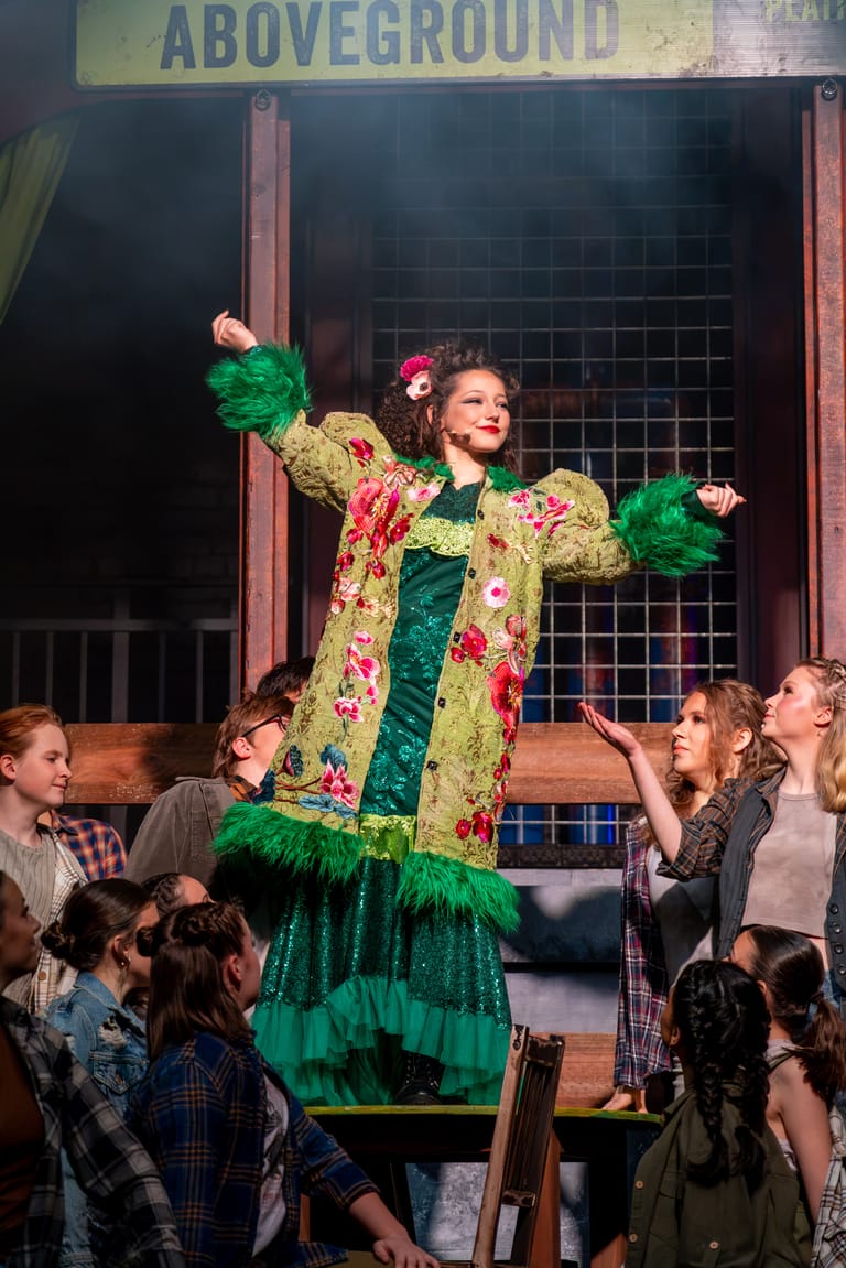 Teen performer in gold and green patterned coat strikes an exuberant pose on stage surrounded by ensemble cast members during Hadestown production.