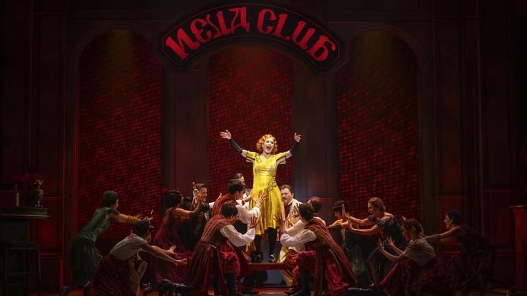 Performer in a gold sequined dress stands centre stage with arms raised, surrounded by dancers in burgundy costumes beneath a red "Moulin Club" sign.