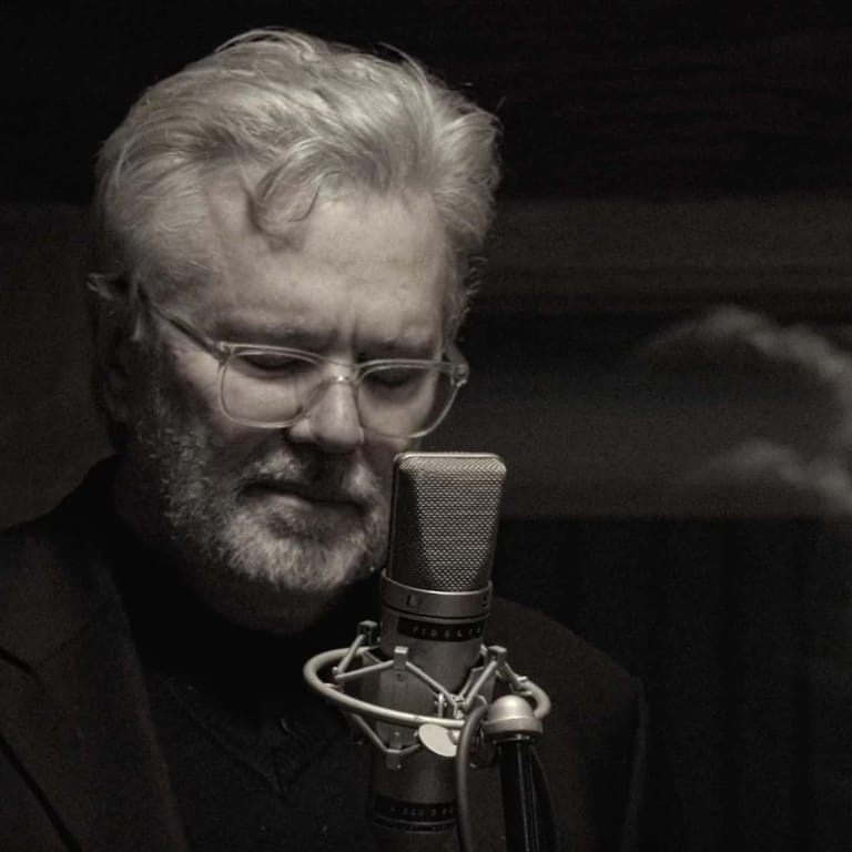 Bearded man wearing glasses singing into a vintage microphone against a dark background.