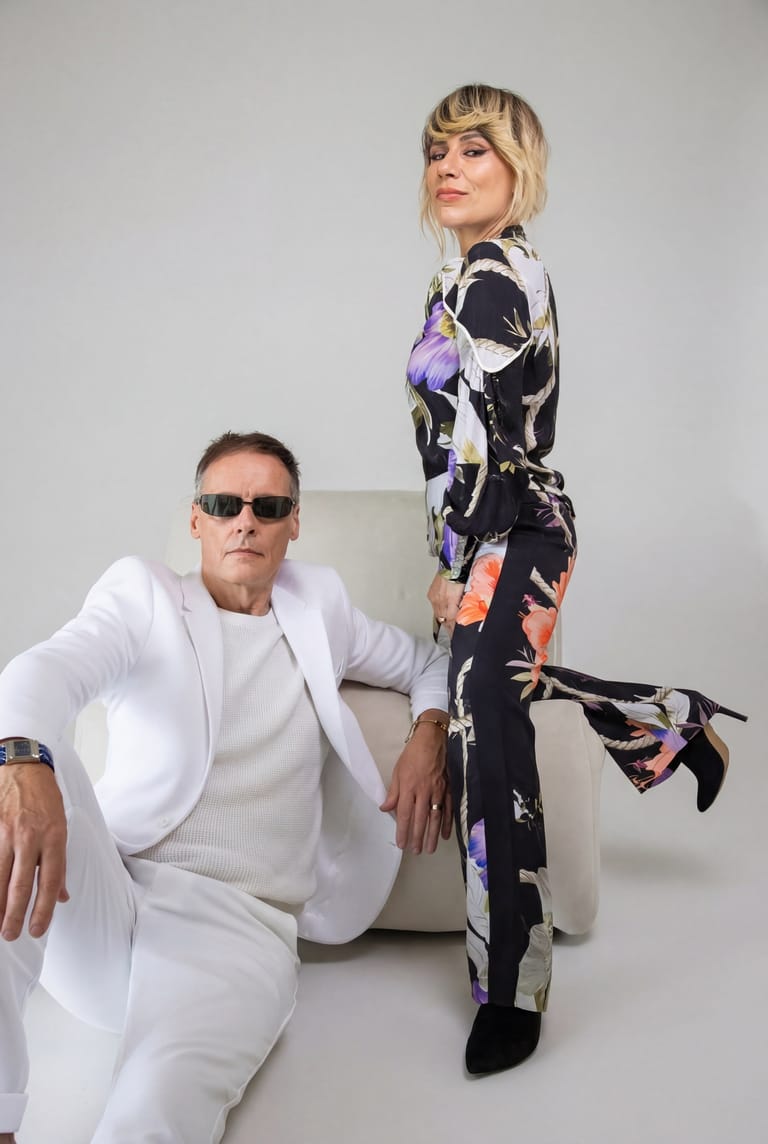 Two people in stylised outfits pose together against a neutral white background; one seated wearing all white, the other standing in a black and purple patterned suit.