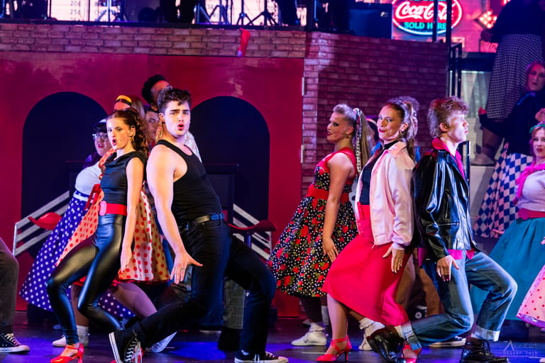 Ensemble cast in 1950s costume dancing on a neon-lit stage set during a Grease theatre production.