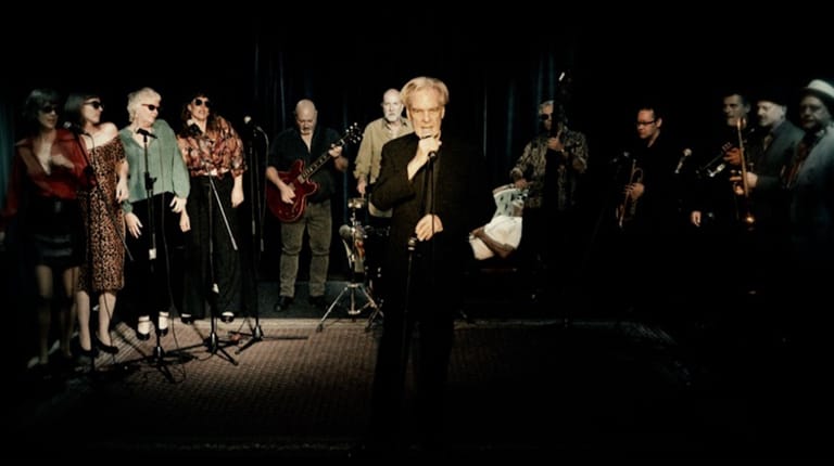 Older man in light-coloured suit speaks at microphone whilst formally dressed audience members stand around him on a darkened stage.