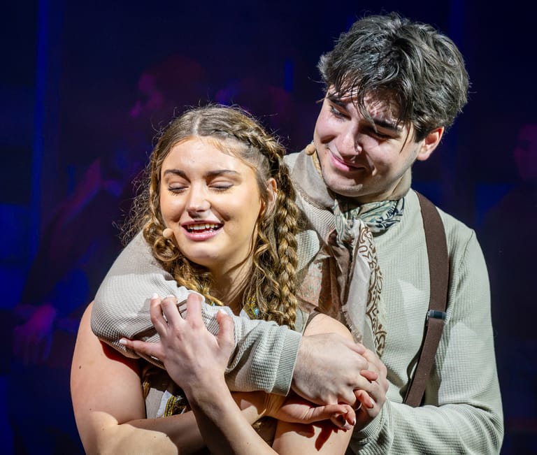 Two performers embrace on stage, lit by blue and purple theatre lighting.