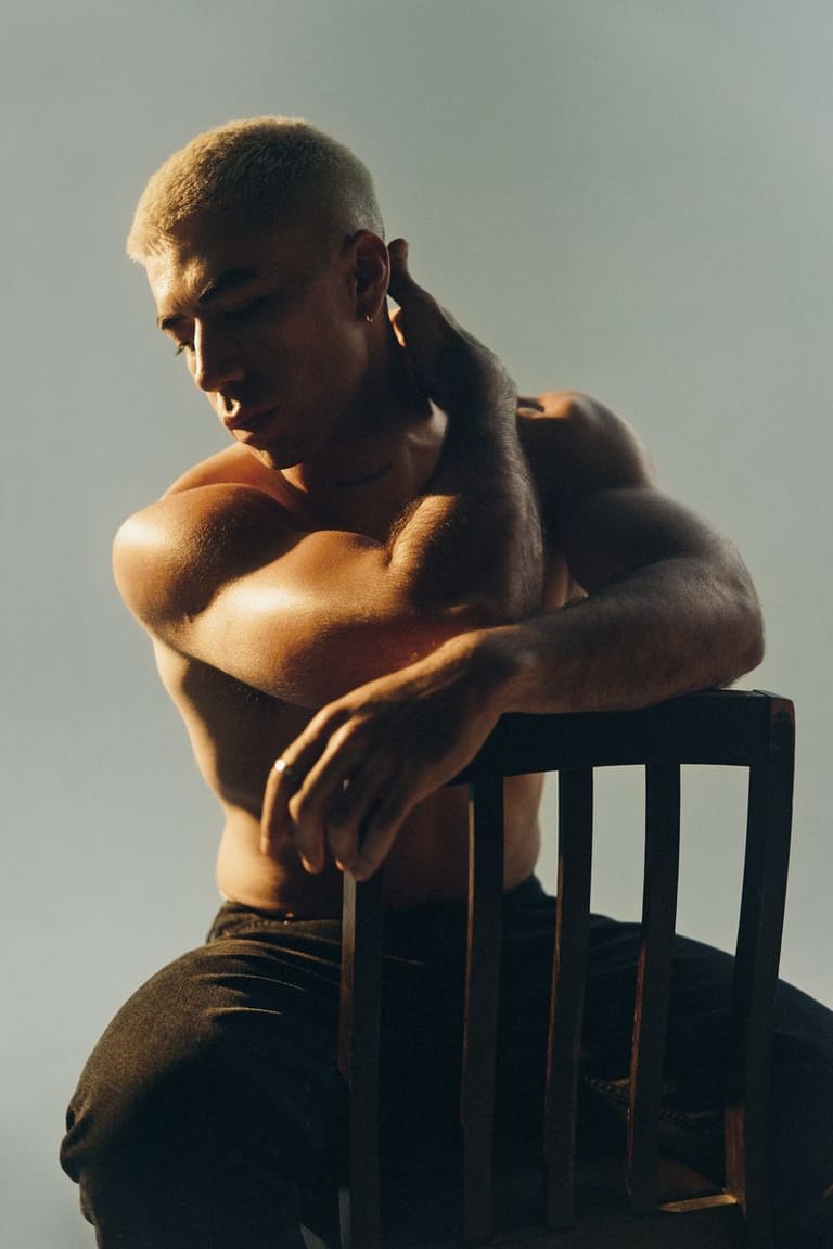 Shirtless man seated on a wooden chair in contemplative pose against a grey background.