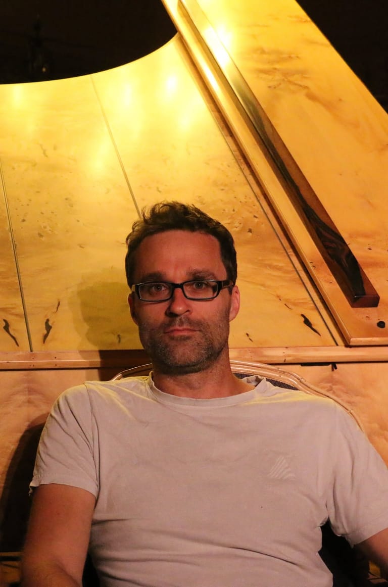 Man wearing glasses and light grey t-shirt posed against warm-lit tent interior.