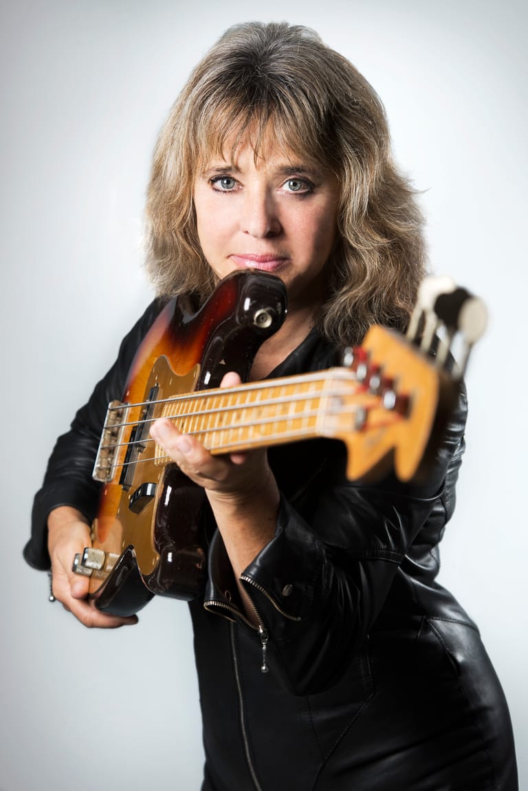 Musician with blonde wavy hair holding an electric guitar, wearing a black leather jacket against a neutral background.