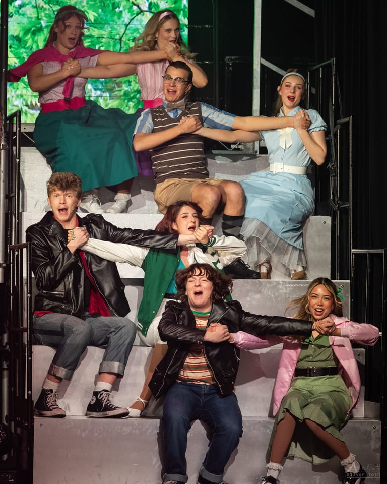Cast members in 1950s-style costumes strike theatrical poses on a bed during a Grease stage production.