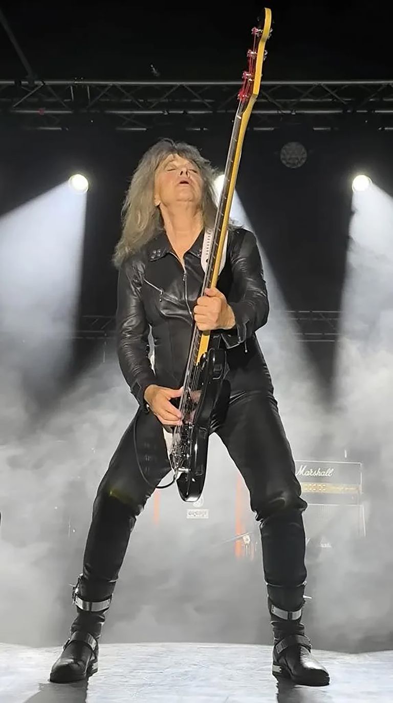 Guitarist in black leather performing on stage with raised instrument, illuminated by dramatic white spotlights and stage fog.