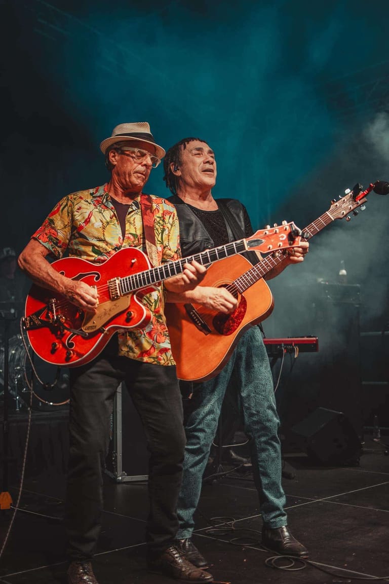 Two guitarists perform on stage against a teal-lit backdrop, one in a patterned shirt with electric guitar, the other with acoustic guitar.