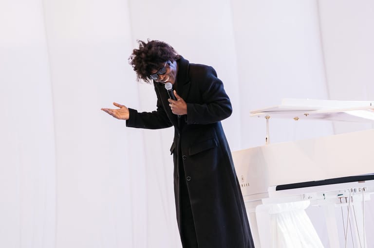 Person with curly dark hair in black coat gesturing expressively against white backdrop.