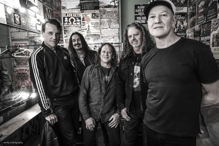 Five musicians in dark clothing stand together against a wall covered with band posters and memorabilia.