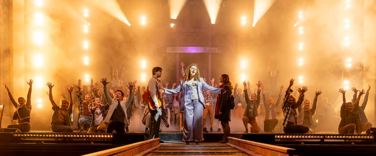 Ensemble cast with arms raised performs on an illuminated stage surrounded by warm golden lighting.