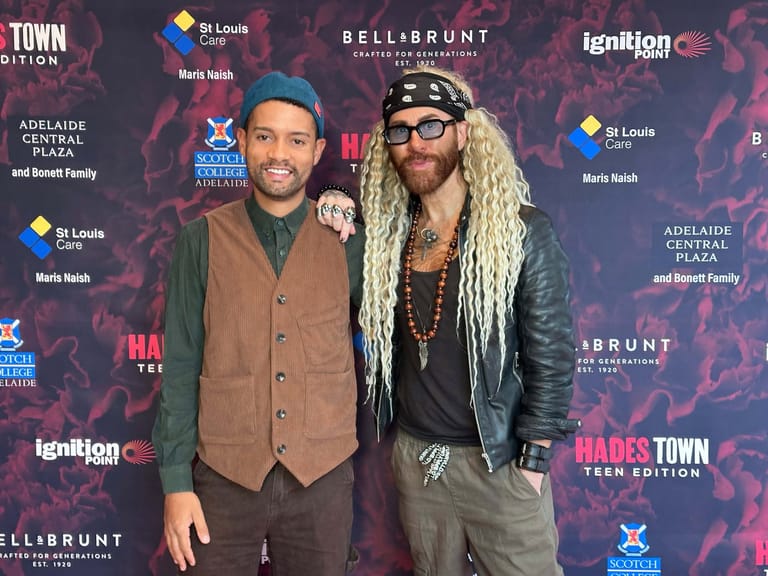 Two men pose together at a media event with branded backdrop, one wearing a brown vest and the other in a leather jacket with long blonde braids.