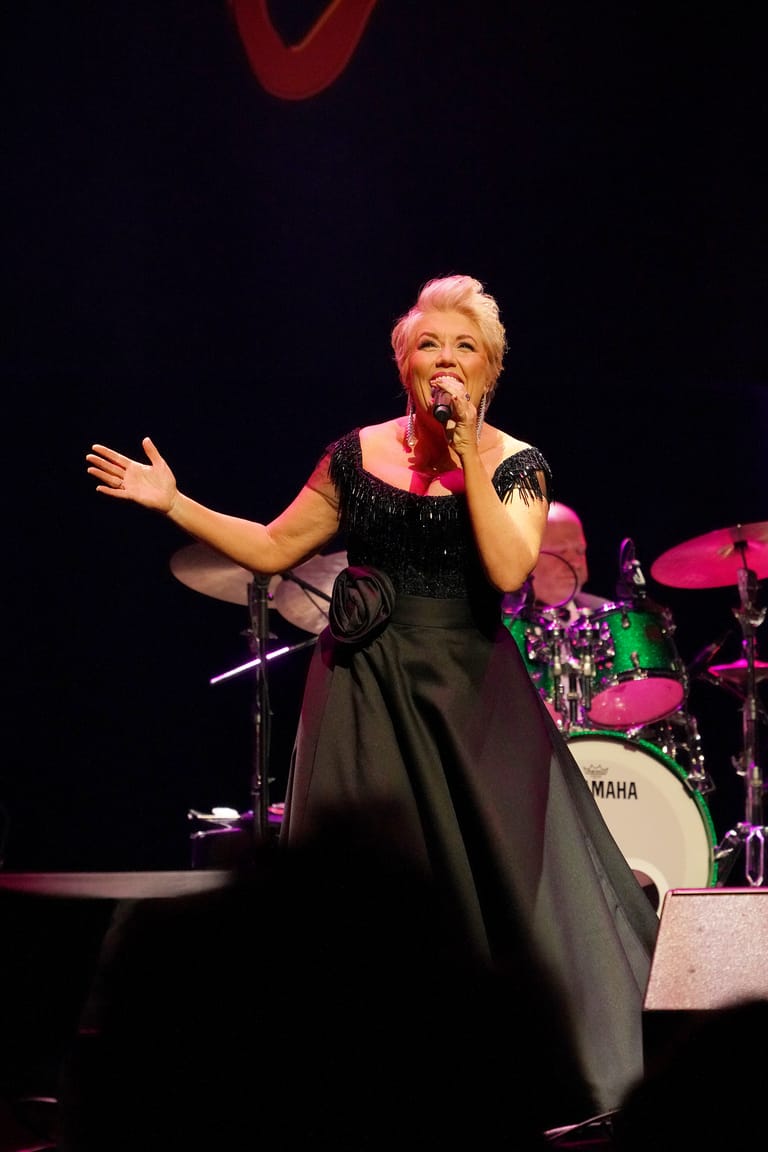 Performer in a black dress and microphone with outstretched arm on a dimly lit stage.