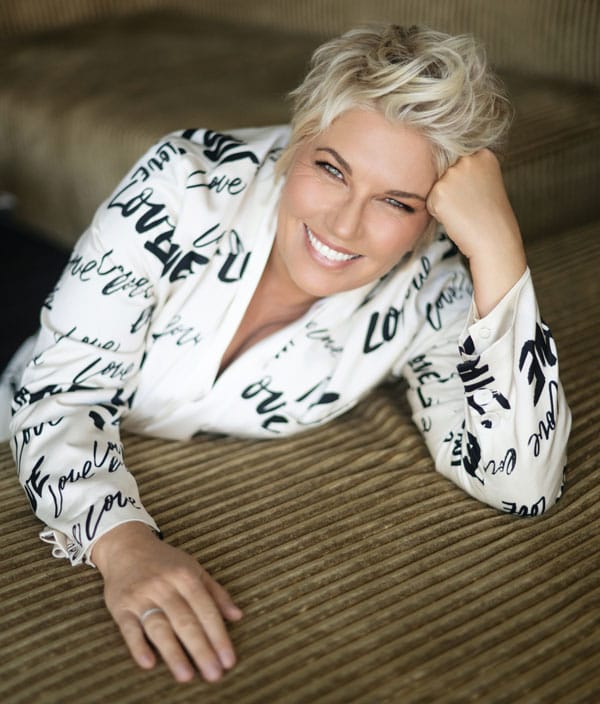 Person with short blonde hair wearing a white patterned jacket, leaning against a striped surface with a warm smile.