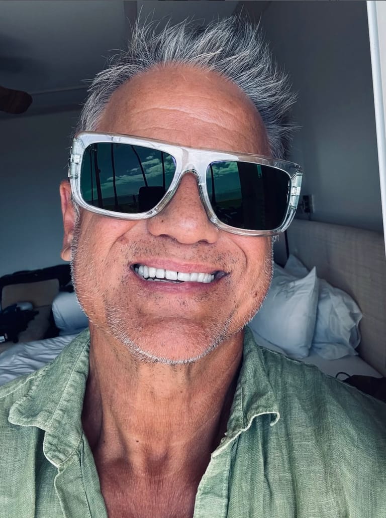 Man wearing white-framed sunglasses and green shirt smiles at the camera indoors.