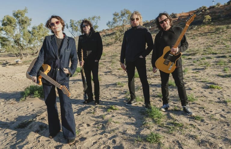 Four musicians with guitars stand on arid ground with sparse vegetation and a hillside backdrop.