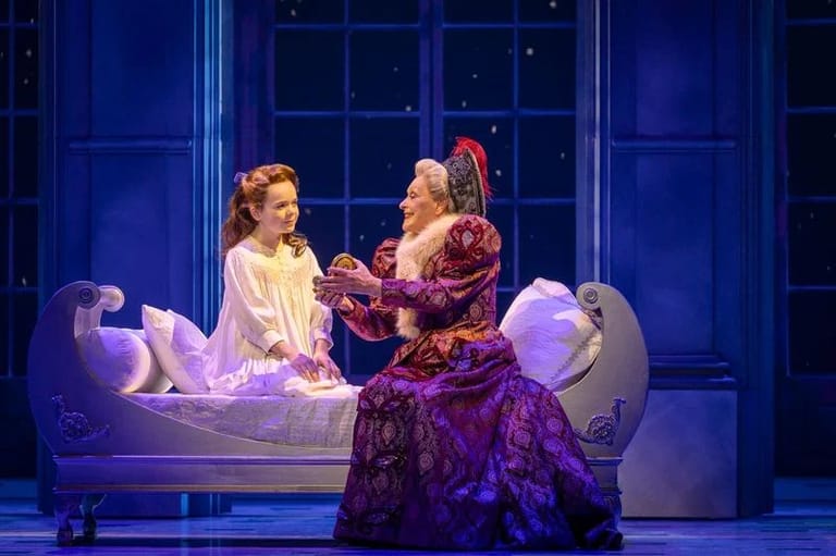Two performers in period costume on stage beside a bed, lit by purple and blue stage lighting.