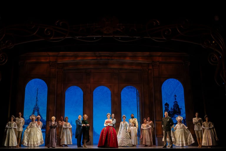 Cast of performers in period costumes standing in a line across a stage beneath ornate arches lit by blue backdrop lighting.