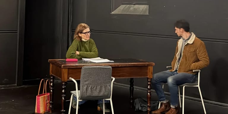 Two people seated at a wooden table during an intimate theatre scene or rehearsal.