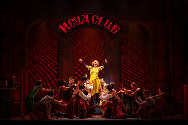 Performer in a yellow dress stands centre stage surrounded by seated dancers in red at the Moulin Rouge theatre.