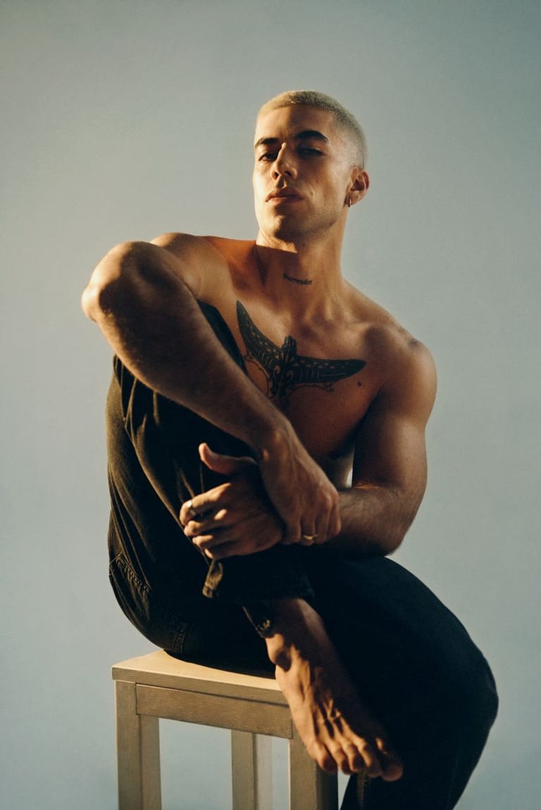 Shirtless man seated on a wooden stool against a pale blue background, gazing thoughtfully to the side.